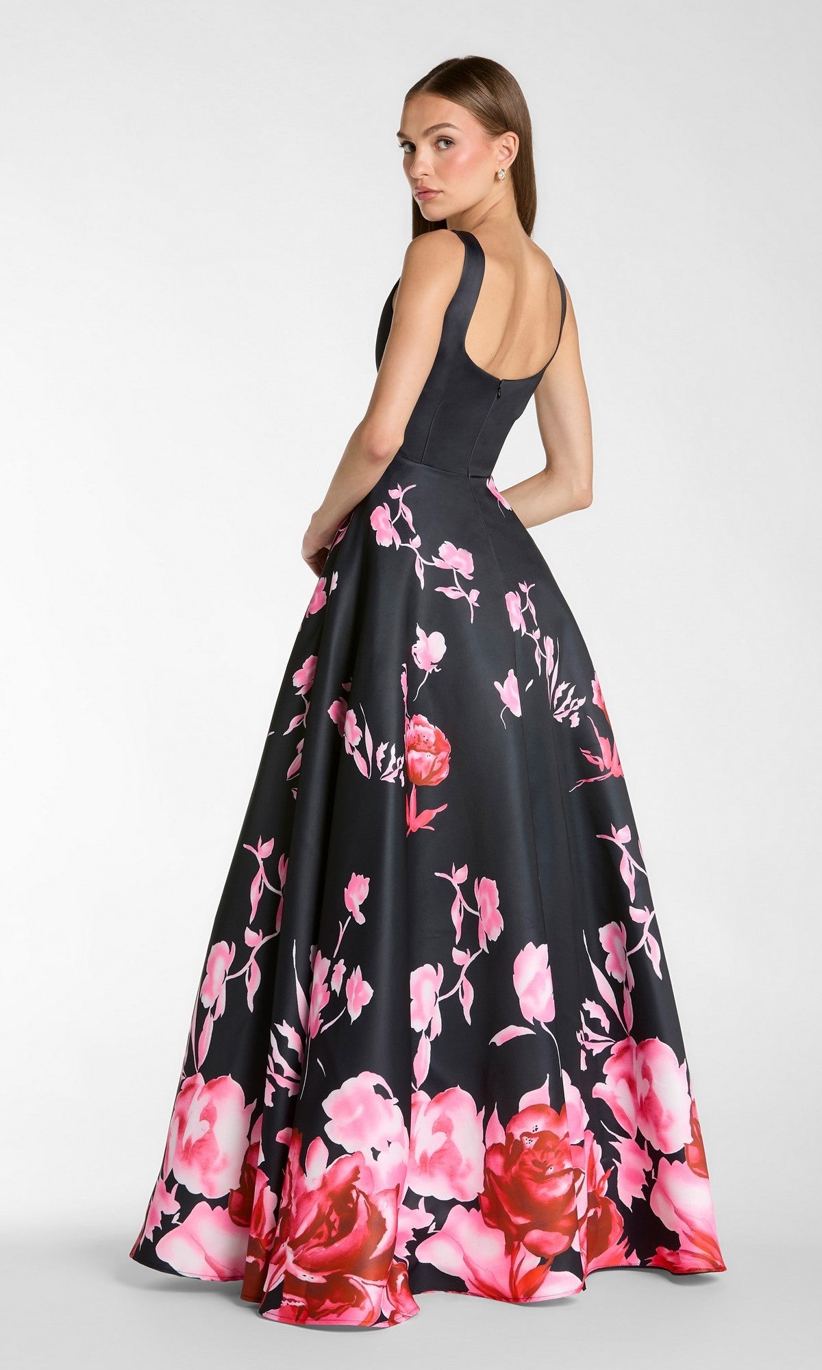 A woman wearing the Mac Duggal 37149 long formal dress—a sleeveless black satin ballgown adorned with a pink floral pattern—stands on a plain background, looking over her shoulder.